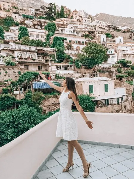Brunette woman in a white dress with arms up toward Positano, Italy ledge pretending to be France Mayes in Under The Tuscan Sun