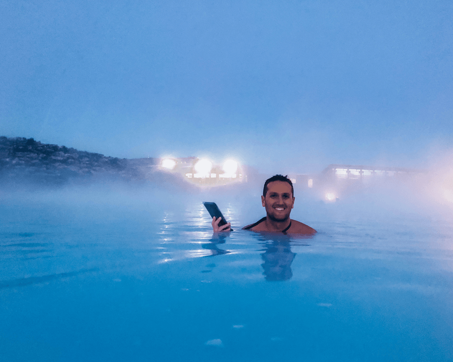 Brunette male in bathing suit in blue waters of Blue Lagoon Geothermal Spa Iceland holding a phone protector
