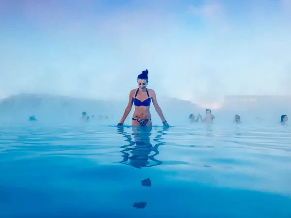 Blue Lagoon In Iceland with white brunette female in a blue bikini with steam and people around her