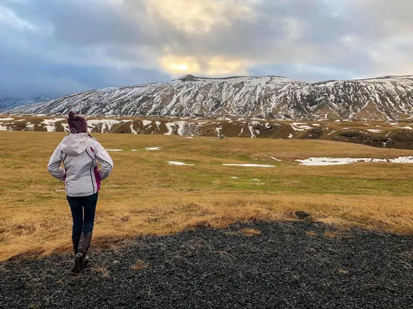 Week In Iceland Hella Road Trip Stop with brunette white woman in a white winter coat standing on  green grass looking at a snow covered mountain