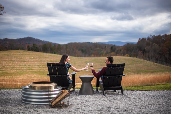 Reasons To Move To Asheville Wineries with brunette white male and female in chairs looking out over Marked Tree Vineyards doing a cheers with wine glasses