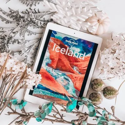 bookstagram flat lay example two with ereader showing a Lonely Planet Iceland travel guide, white and turquoise flowers, and tree candles