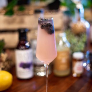 Blackberry Smash Cocktail champagne glass filled with pink drink and vodka with floating blackberries in front of a blurred backdrop of cocktail ingredients