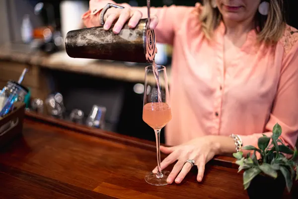 How to make a blackberry smash with woman in a pink blouse pouring pink liquid into a glass from a shaker with a strainer on a bar top