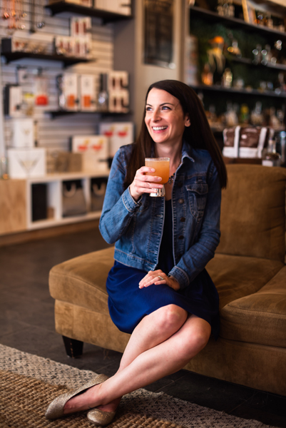 White, brunette woman drinking a peach and gin cocktail on a brown sofa at Cultivated Cocktails in Asheville, NC