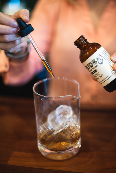 How To Make A Smokey Old Fashioned Cocktail with woman in pink shirt dropping bitters into a cocktail mixing glass with ice cubes and whiskey