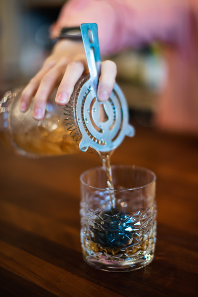 How to make a smokey old fashioned cocktail picture with woman in pink shirt pouring a strained smoked old fashioned into an old fashioned low ball glass with a whiskey ball