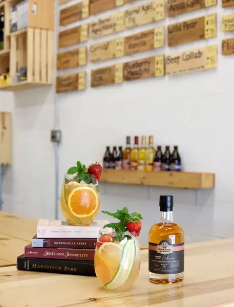 Lady Jane Literary Mead Cocktail with two cocktails, stack of Jane Austen books, and bottle of mead with meadery taproom in background
