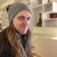 Jeremy Paterson Bio picture with white male with long redish-brown hair wearing a gray hat and dark shirt in front of laptop with bookshelf in the background