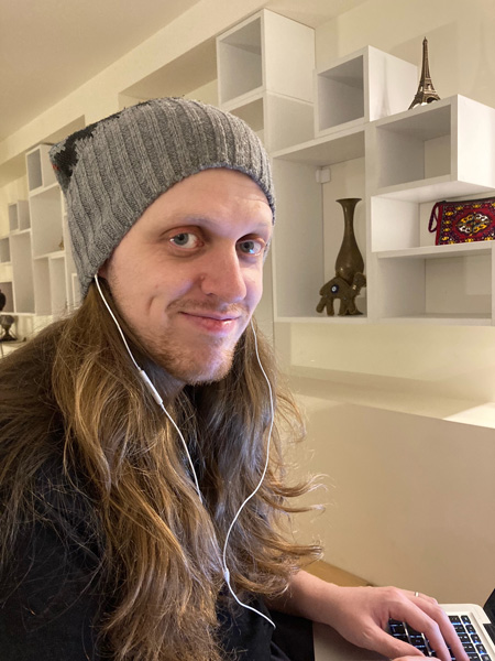 Jeremy Paterson Bio picture with white male with long redish-brown hair wearing a gray hat and dark shirt in front of laptop with bookshelf in the background