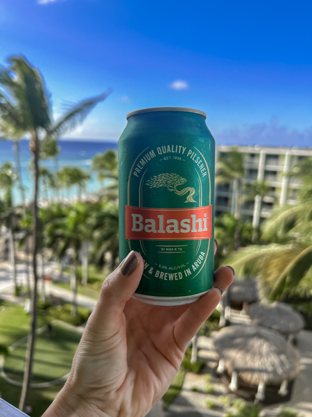 Balashi Beer in Aruba with white hand holding up a green can of beer with palm trees, resort, and beach in the background