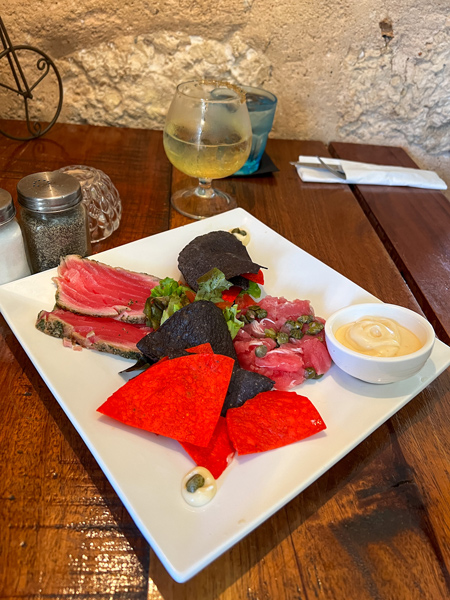 Bistro de Suikertuin in Aruba seared raw tuna and raw tuna chunks on white plate with red and purple tortilla chips next to yellow cocktail on brown table