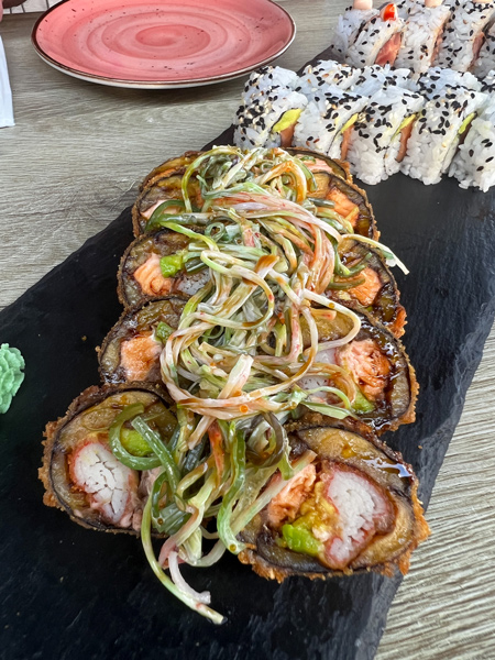 Nikkei Sushi Wok & Bowl Restaurant in Aruba with image of sushi roll filled with crab, salmon, and plantains on black plate