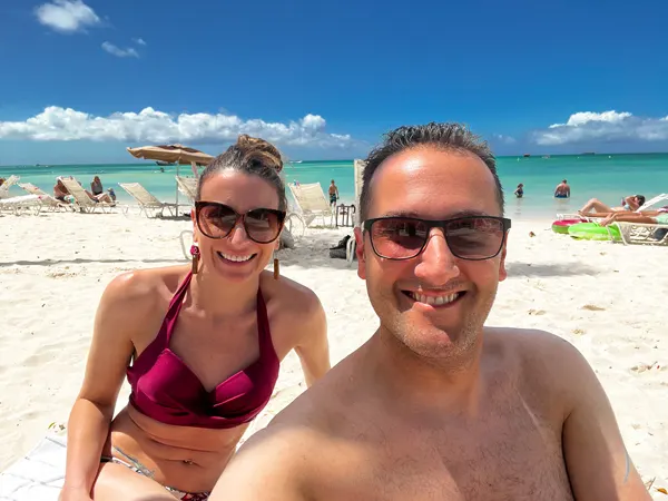 Palm Beach in Aruba with white brunette male and female in sunglasses and bathing suits on white-ish sand with blue water in the background