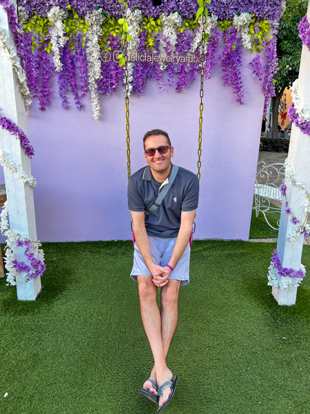 Paseo Herencia Mall Instagram Swing with white brunette male wearing sunglasses, t-shirt, and shorts with purple flower backdrop