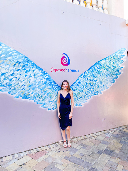 Paseo Herencia Mall blue wing mural with white brunette woman standing in front of it wearing a black dress and sandals