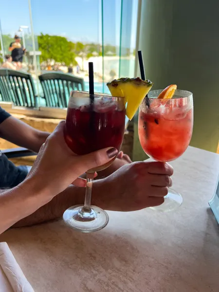 Rum Reef Cocktail Bar & Grill in Aruba with two white hands doing a cheers with red sangria and watermelon cocktail in wine like glasses with pool blurred in background