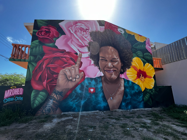 Sarah Quita Mural in San Nicolas, Aruba with woman with finger showing number one symbol surrounded by red, yellow, and pink flowers