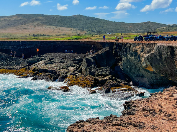 UTVs around Natural Cave Pool in Aruba with bright blue water, green grass, and ladder going down to the cave pool