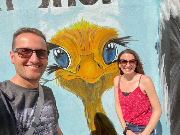 Aruba Ostrich Farm Entrance with mural of yellow orange ostrich face and white brunette male and female in sun glasses taking a selfie