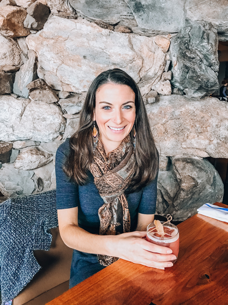 Christine Frascarelli, a white brunette female wearing a gray dress and sitting at a table with a pink cocktail and a stone wall behind her